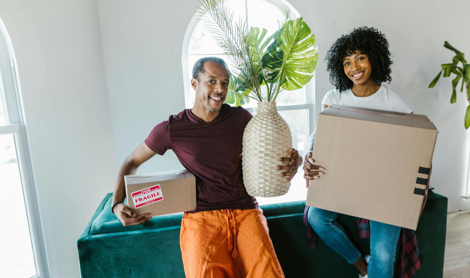 Couple smiling while moving into a new home, carrying boxes and a plant.