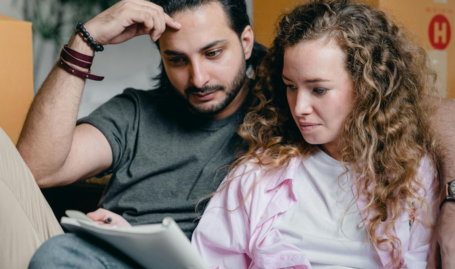 Focused male and female in casual wear sitting near boxes and looking through notebook thoughtfully while moving house